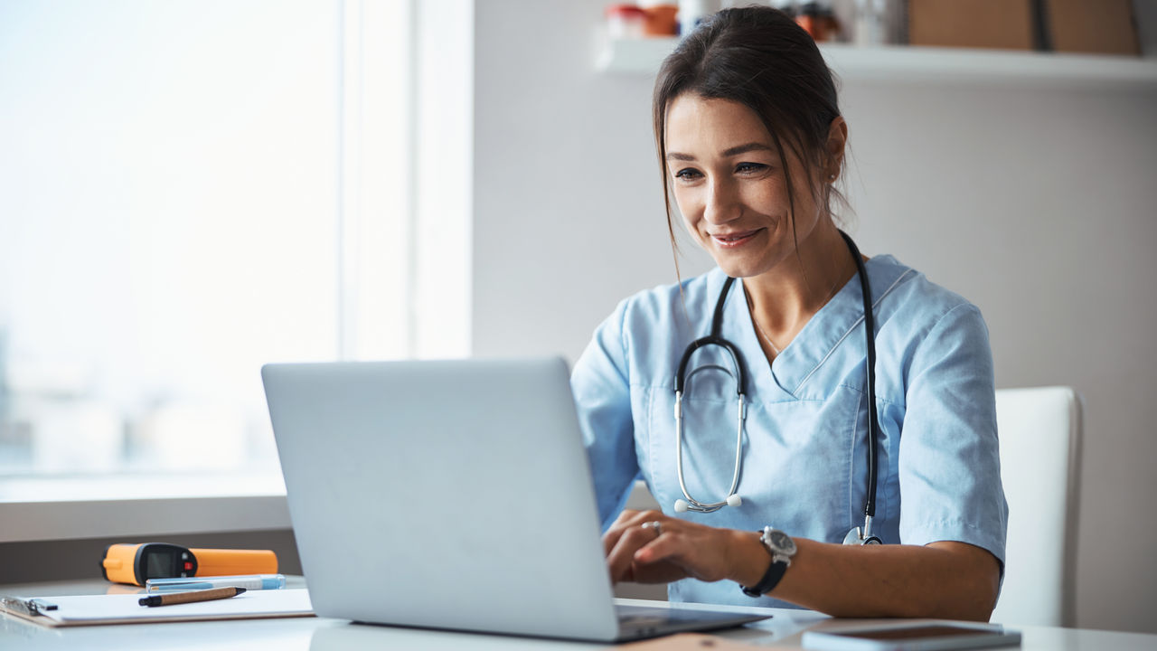 Beautiful young woman physician sitting at the table and smiling while working on modern laptop
