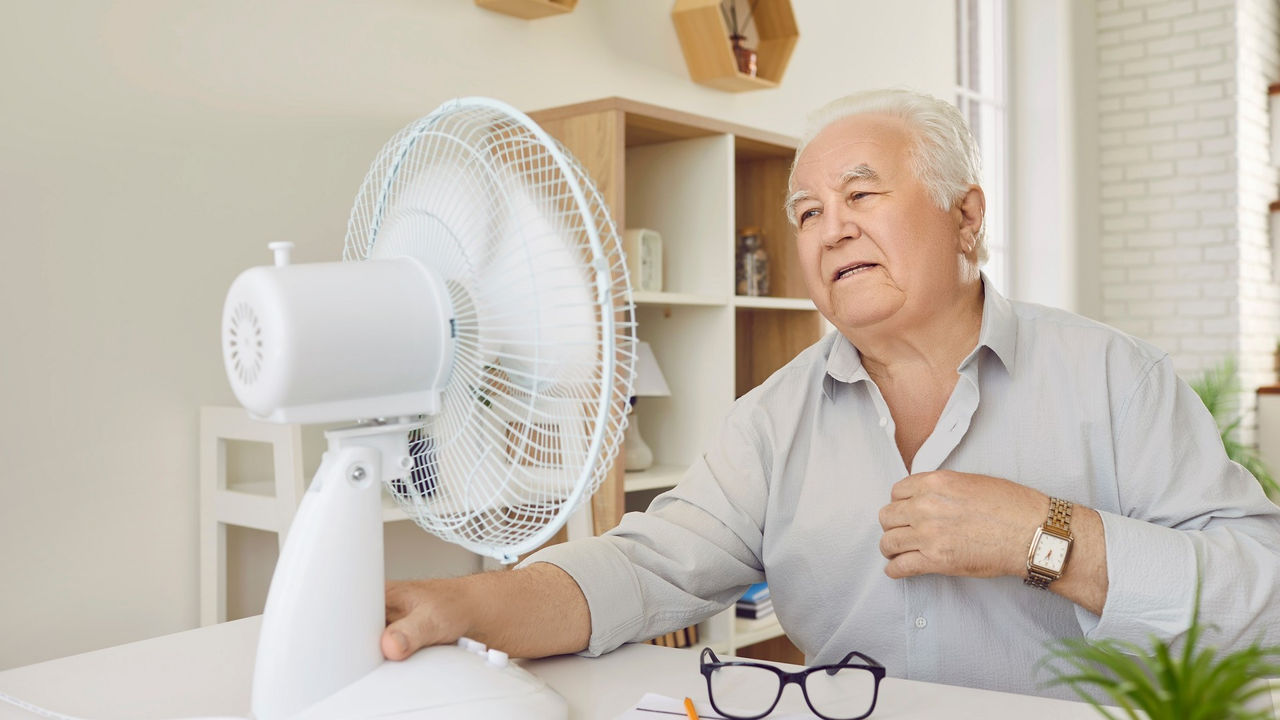 Tired upset senior retired person using electric fan during heatwave sitting at the desk.